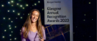 A woman stands holding a trophy before a sign advertising the Glasgow Annual Recognition awards