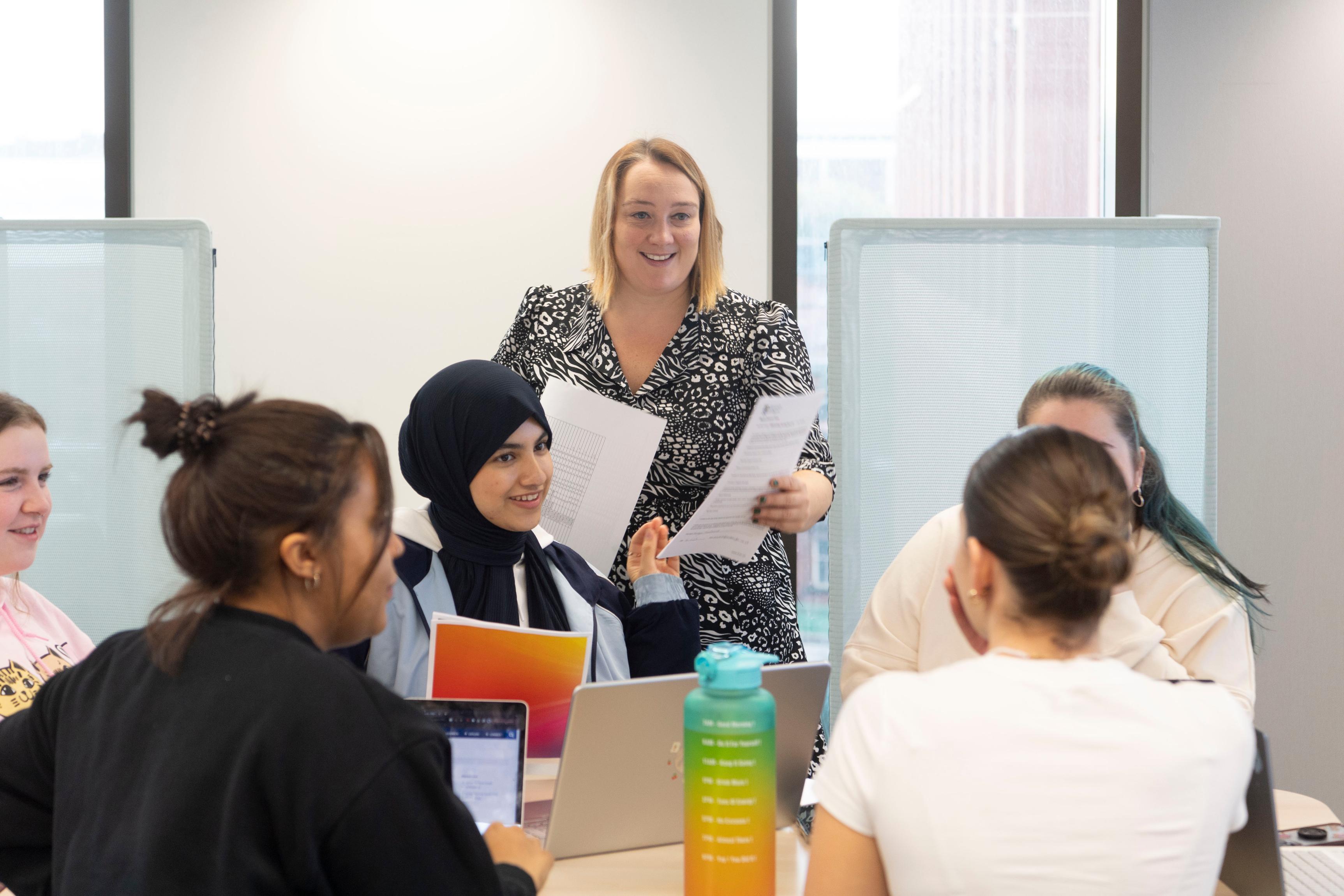 A blonde woman stands before a table of students, who are all smiling as she hands out worksheets