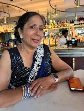 Photo of Mrs Pinky Virhia - she is smiling while sitting at a table, with her hands clasped on top of the table in front of her.  She is wearing a blue and white outfit and long sparkling earring in her ears