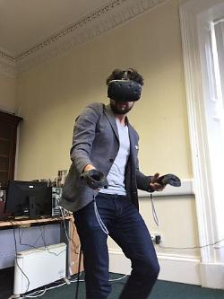 photo of a man in jeans, a blazer and white tshirt, with black beard and curly hear, kitted out with a virtual reality headset and two handheld controllers, moving inside a classroom. in the background there are wires and a computer set up