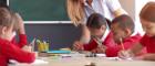 An image of children in a classroom writing on a desk with a teacher leaning in to help support